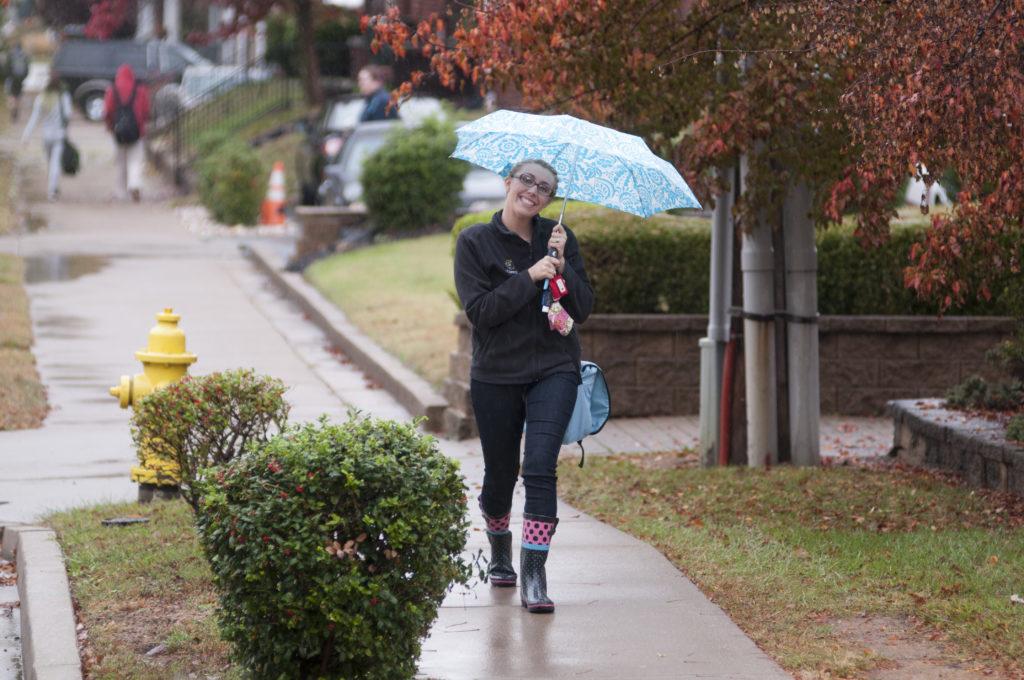 Student walking in the rain, with an umbrella, smiling at the camera.